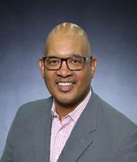 A man with glasses, a bald head, and a trimmed beard, smiling and wearing a gray blazer over a pink checkered shirt, poses in front of a dark gradient background.