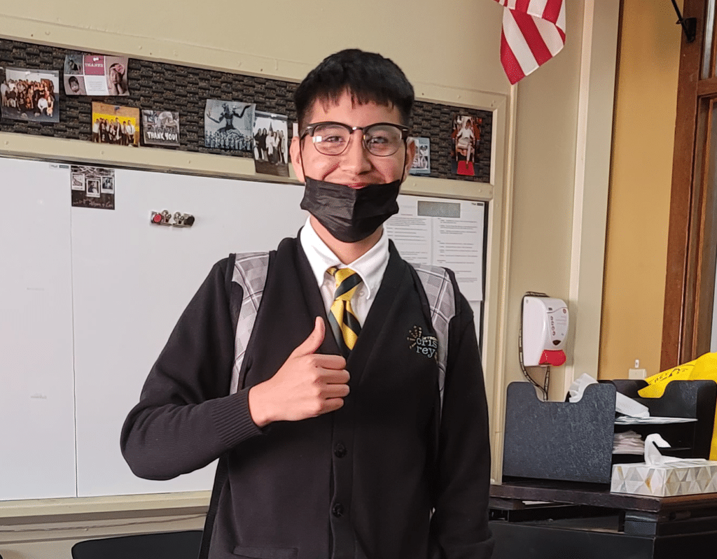 A student wearing glasses, a black mask below his chin, a black cardigan, and a striped tie gives a thumbs-up in a classroom with photos, a whiteboard, and an American flag in the background.