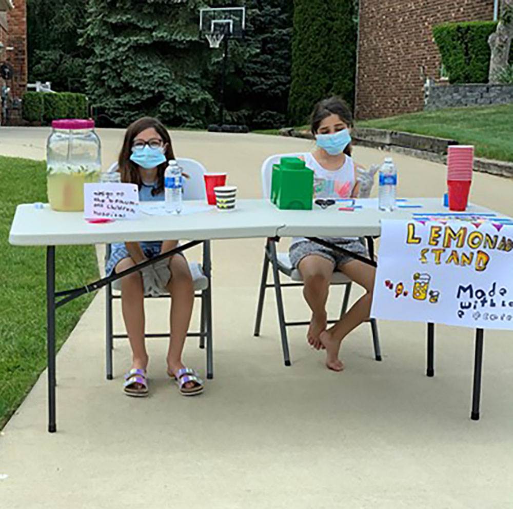Two children wearing masks sit behind a table on a driveway, selling lemonade. The table has lemonade, cups, water bottles, and a sign that reads "Lemonade Stand" with hand-drawn decorations.