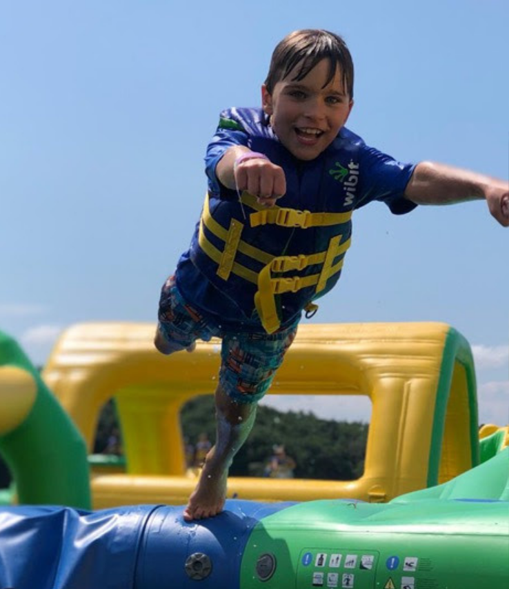 A smiling child wearing a blue life jacket and plaid swim trunks leaps mid-air on an inflatable water obstacle course under a sunny sky.
