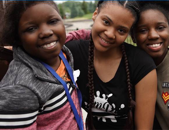 Three smiling girls stand close together outdoors, posing for the camera. The girl on the left wears a gray and pink jacket, the middle girl has long braids and a black shirt, and the girl on the right wears a brown jacket.
