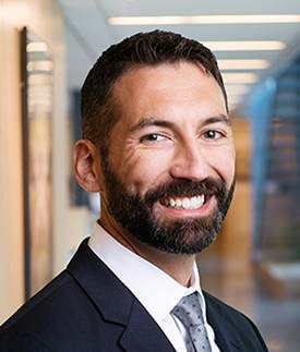 A man with short dark hair and a beard, wearing a dark suit, white shirt, and gray tie, smiles at the camera. The background is an office hallway with soft lighting and blurred details.