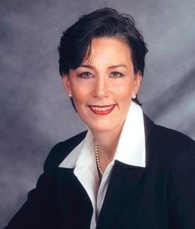 A woman with short dark hair, wearing a black blazer, white collared shirt, and a pearl necklace, smiles at the camera against a gray textured background.