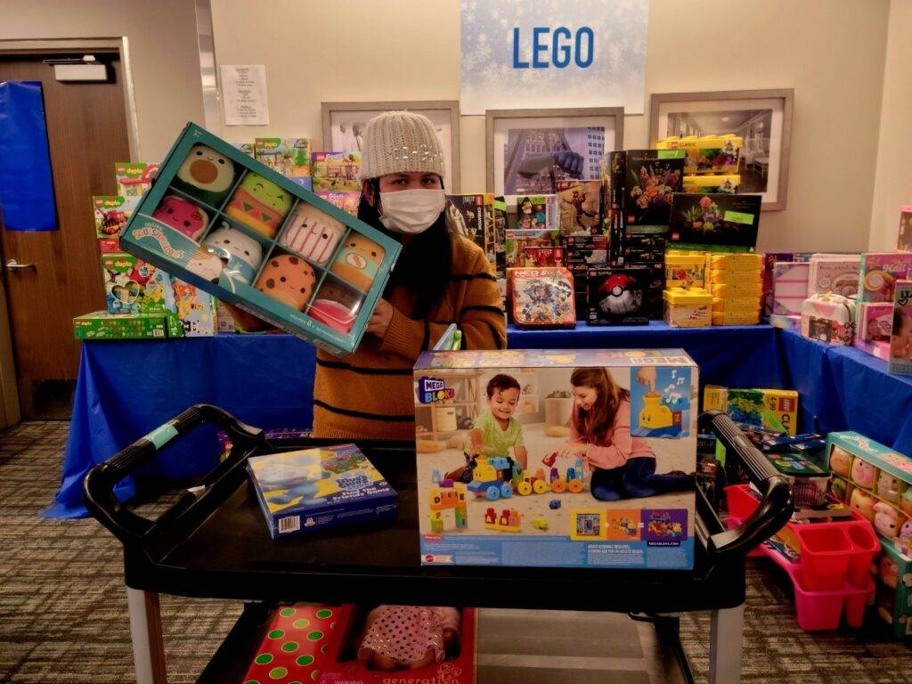A person wearing a beanie and mask holds up a toy set in front of a display of colorful toys, with a LEGO sign above. More toys are arranged on tables and on a cart in the foreground.