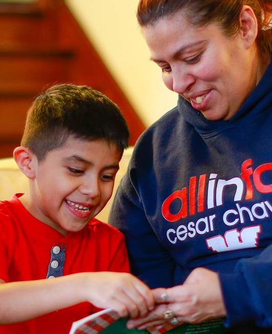 A smiling woman and a boy sit closely together, looking at and pointing to something in a book. The woman wears a dark sweatshirt, and the boy wears a red shirt. They appear happy and engaged in the activity.