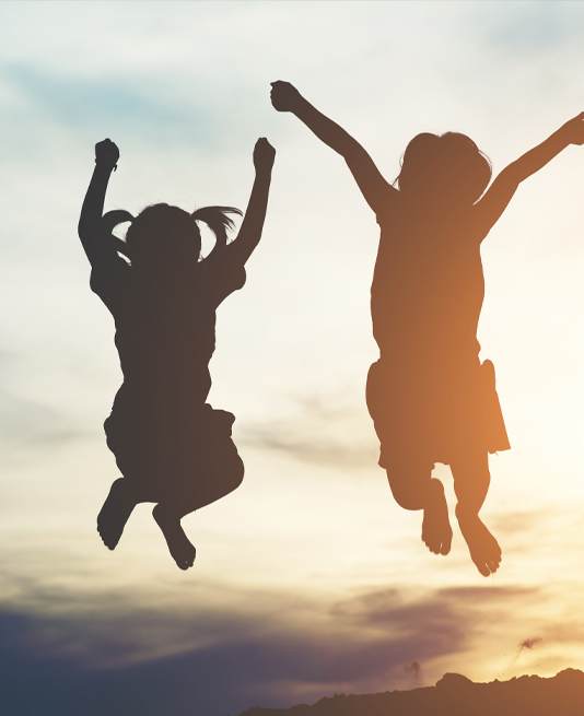 Two children are silhouetted against a sunset sky, jumping with their arms raised in joy.