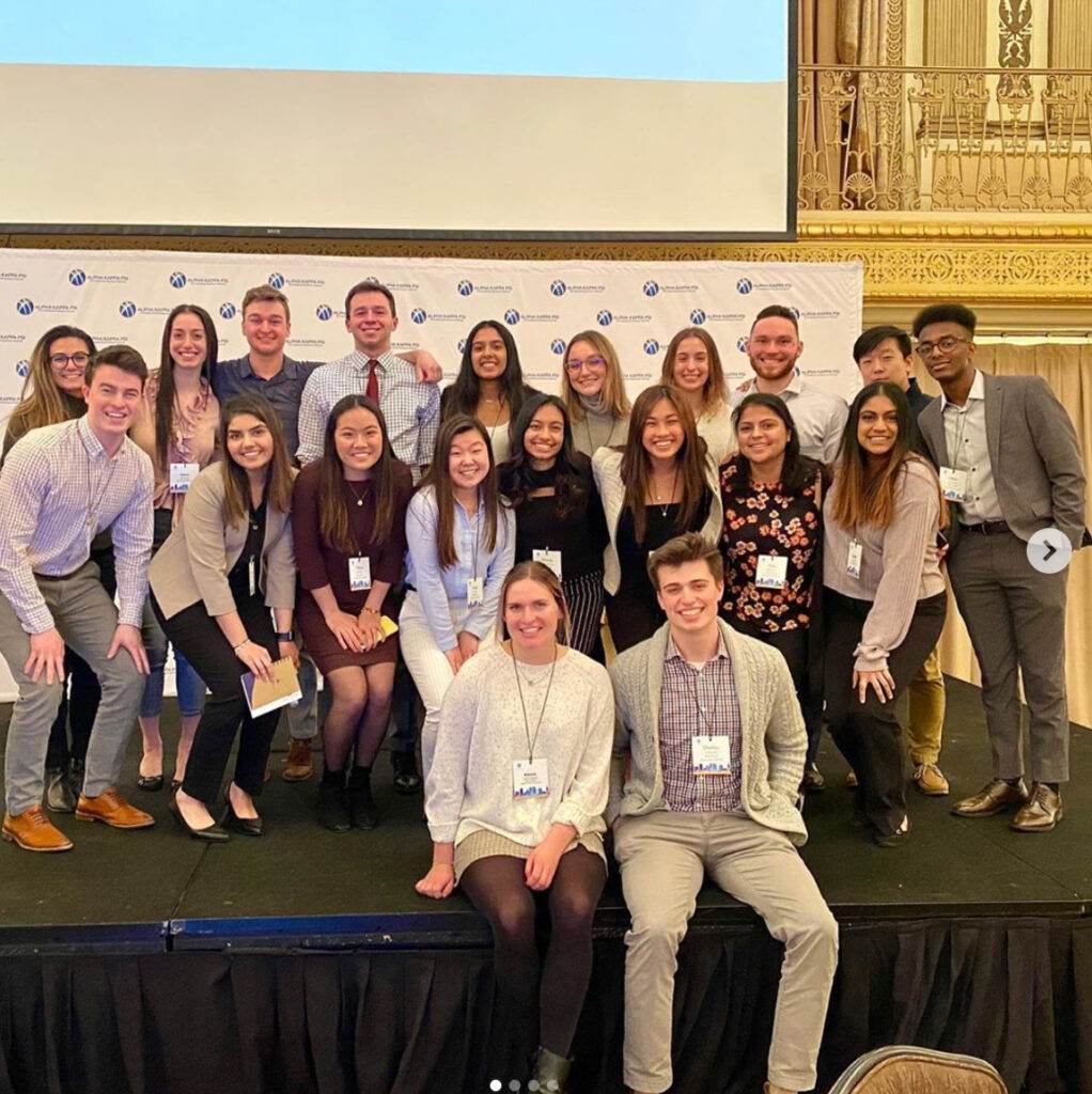 A group of young adults, dressed in business casual attire, smile and pose together on a stage in front of a white banner at a conference or professional event inside an ornate, elegant room.