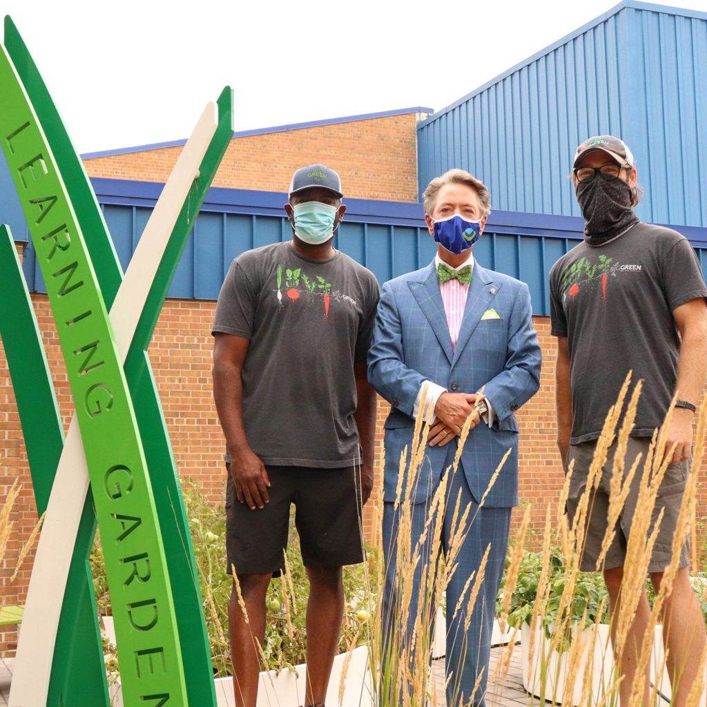 Three men stand in a garden next to a green "Learning Garden" sign. Two wear matching t-shirts, shorts, and face masks; one wears a blue suit and bowtie. A blue and brick building is in the background.