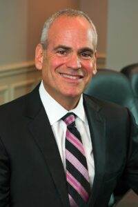 A smiling man with short gray hair wearing a black suit, white shirt, and a pink-striped tie, seated indoors in an office or conference room.