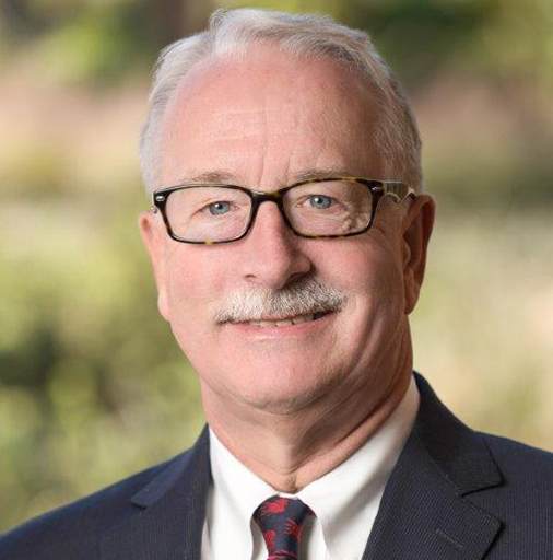 A smiling older man with white hair, glasses, and a mustache is wearing a dark suit, white shirt, and patterned tie. The background is blurred greenery.