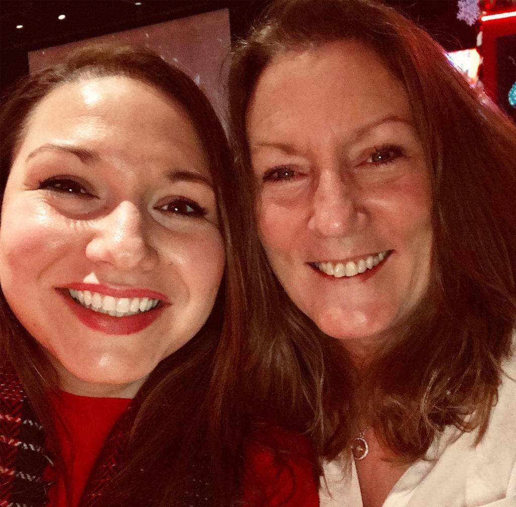 Two women smiling at the camera in a warmly lit indoor setting. Both have long brown hair and are wearing red and white clothing. The background features festive, possibly holiday-themed, decorations.