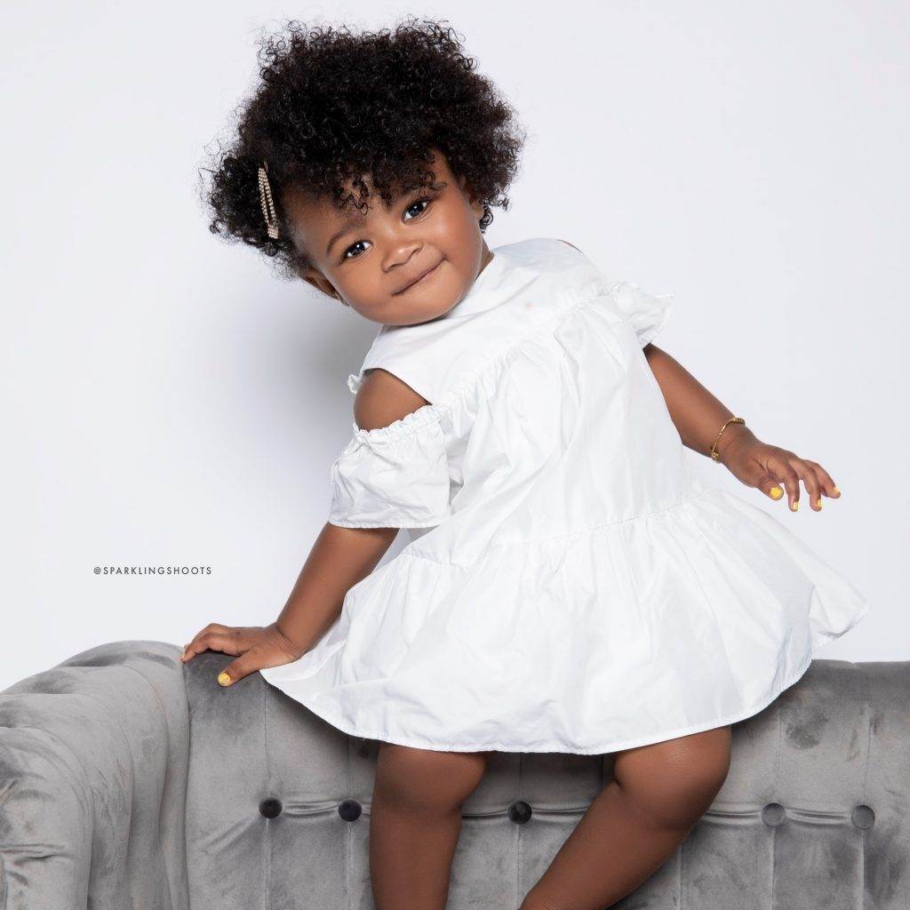 A young child with curly hair wearing a white dress stands on a gray tufted couch, smiling slightly with one hand on the couch back and the other on the seat, against a plain white background.