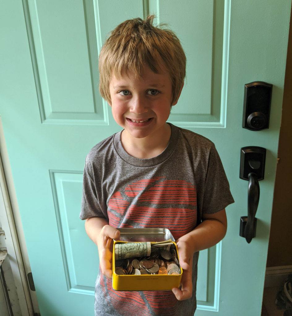 A young boy with light brown hair, wearing a gray and red t-shirt, stands in front of a teal door, smiling and holding a yellow box filled with coins and a rolled-up bill.