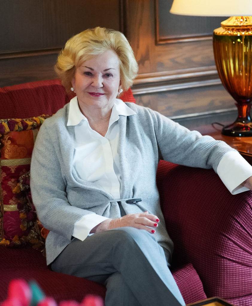 An older woman with short blonde hair, wearing a white blouse and light gray cardigan, sits on a red couch, smiling at the camera. There is a decorative pillow and a lamp in the background.