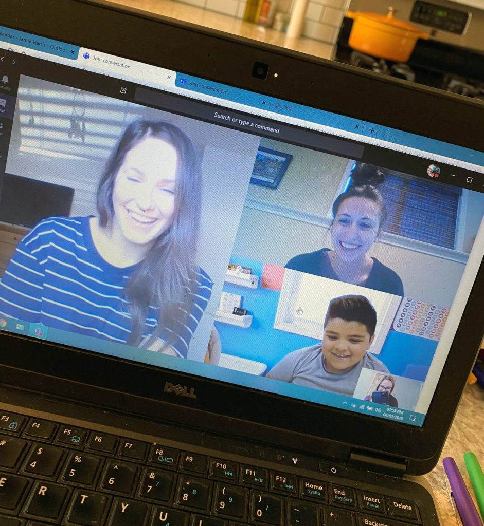 A laptop screen shows a video call with three people: two women and one boy, all smiling. The laptop is on a kitchen counter, with a stove, orange pot, and white tiles visible in the background.