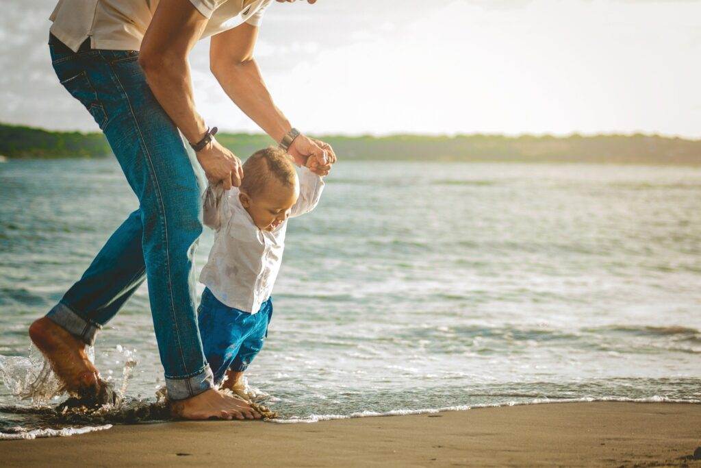 An adult helps a toddler walk along the shoreline, holding the child's hands as small waves touch their bare feet. Both are wearing rolled-up jeans, and the scene is warm and peaceful by the water.