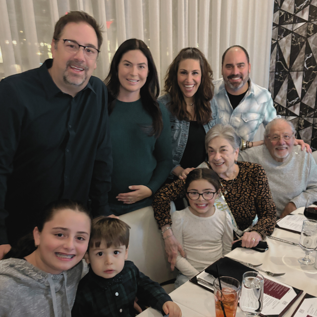 A group of eight people, including adults and children, gather around a dining table at a restaurant, smiling for the camera. The table has drinks, menus, and glasses, and a white curtain hangs in the background.