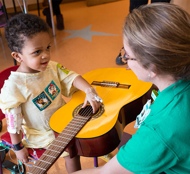 A young child with medical tubes attached touches the strings of a yellow acoustic guitar, guided by an adult woman in green scrubs, in a brightly lit room.