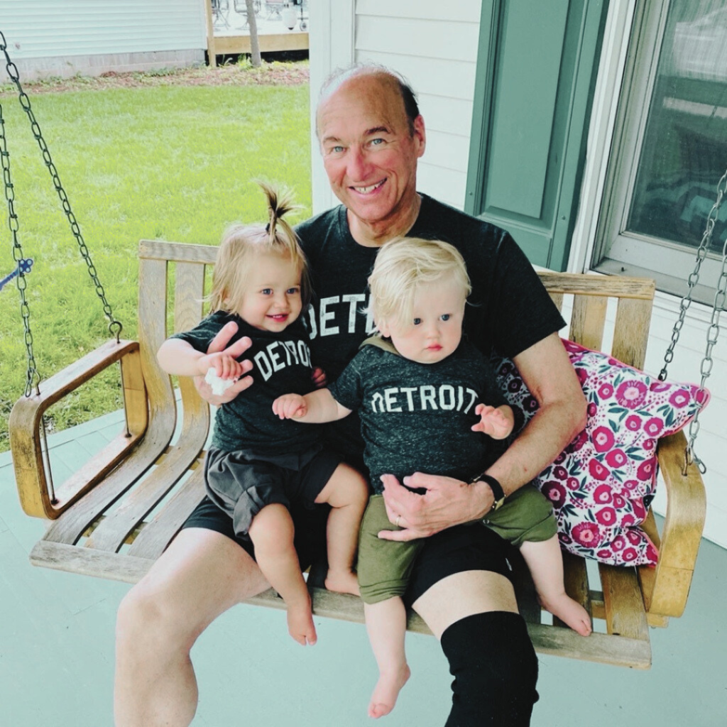 An older man and two small children sit on a porch swing, all wearing matching black "Detroit" shirts. The man holds both children, one of whom is smiling and making a peace sign. A pillow is beside them.