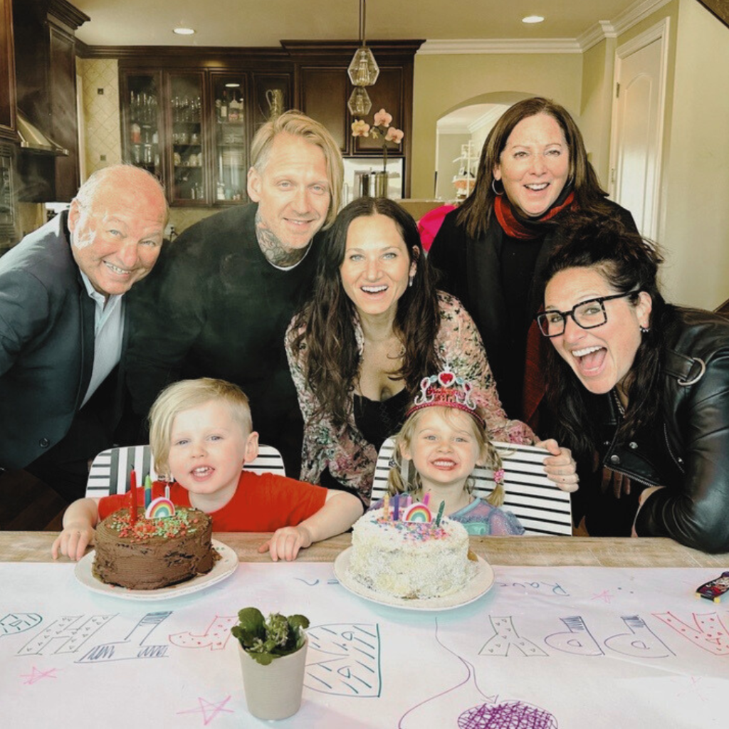 A joyful family gathers around a table with two young children and birthday cakes. The children sit in front, smiling, while the adults stand behind them, also smiling. Colorful decorations and drawings cover the table.