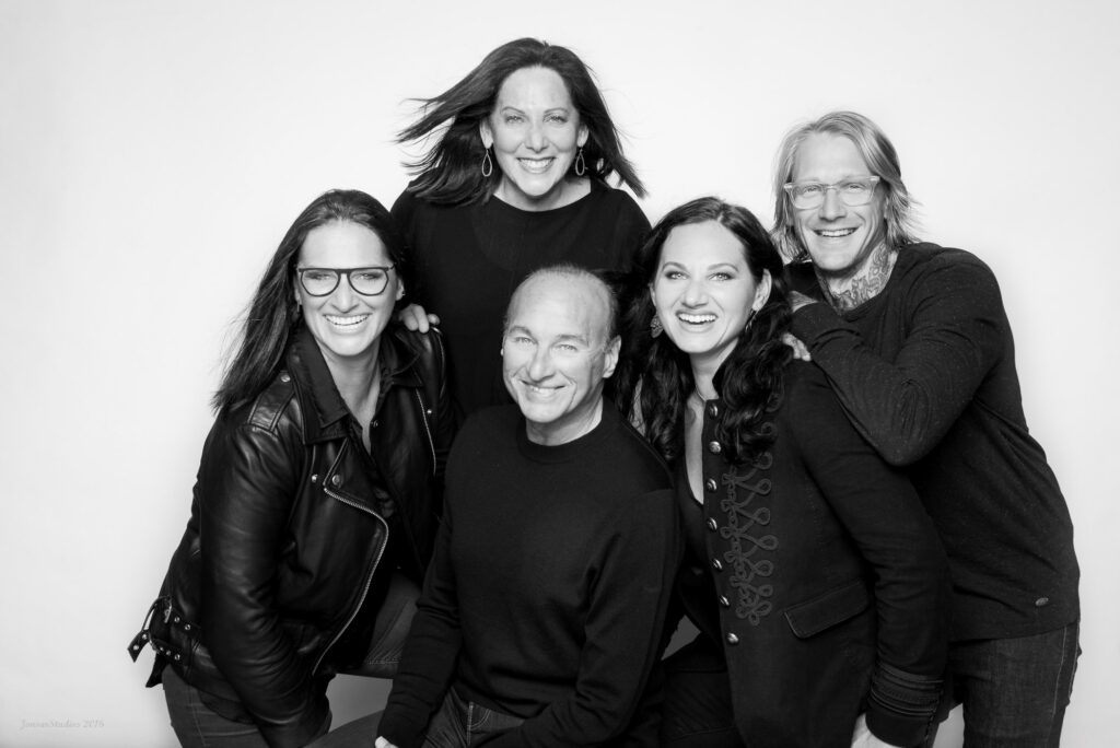 Five adults pose closely together, smiling and laughing, in a black-and-white studio portrait with a plain background. Three women and two men are casually dressed and appear happy and relaxed.