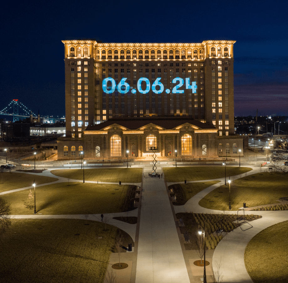 A large, illuminated building at night—Michigan Central Station—displays "06.06.24" in bright blue numbers on its front. Well-lit paths, grass, and trees surround the station, with a cityscape and a bridge in the background.