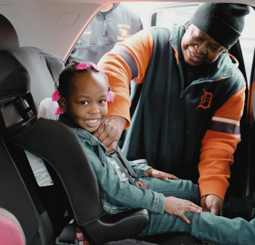 An adult helps a smiling young girl, seated in a car seat, buckle up inside a car. The girl wears a denim jacket and has pink bows in her hair. The adult wears an orange and black jacket and a black beanie.