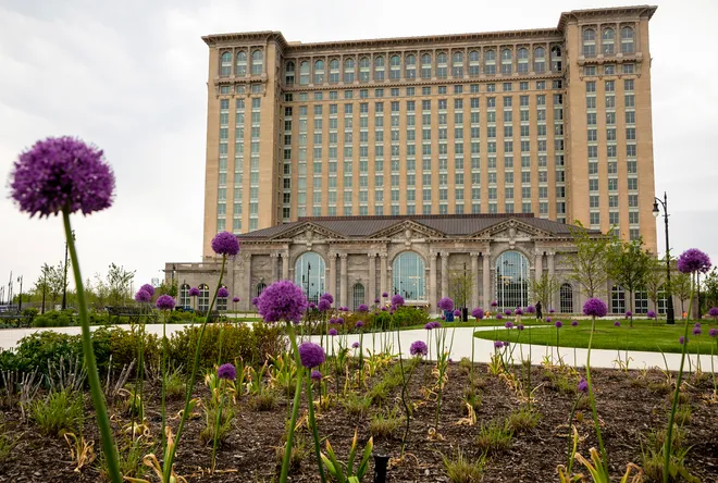 A tall, historic building with many windows stands behind a landscaped garden with blooming purple allium flowers and green shrubs on a cloudy day.
