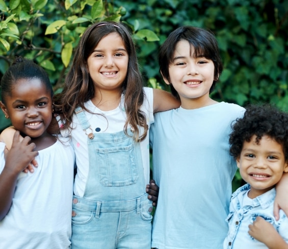 Four smiling children stand close together outdoors, with their arms around each other, in front of green leafy bushes. They look happy and are wearing casual clothes in light colors.
