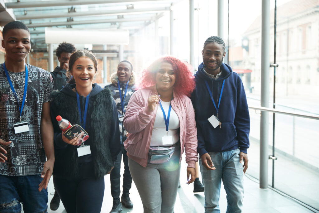 A group of smiling young adults with name badges walk together indoors by large windows. One person holds a bottle, and they appear to be enjoying themselves in a bright, modern setting.