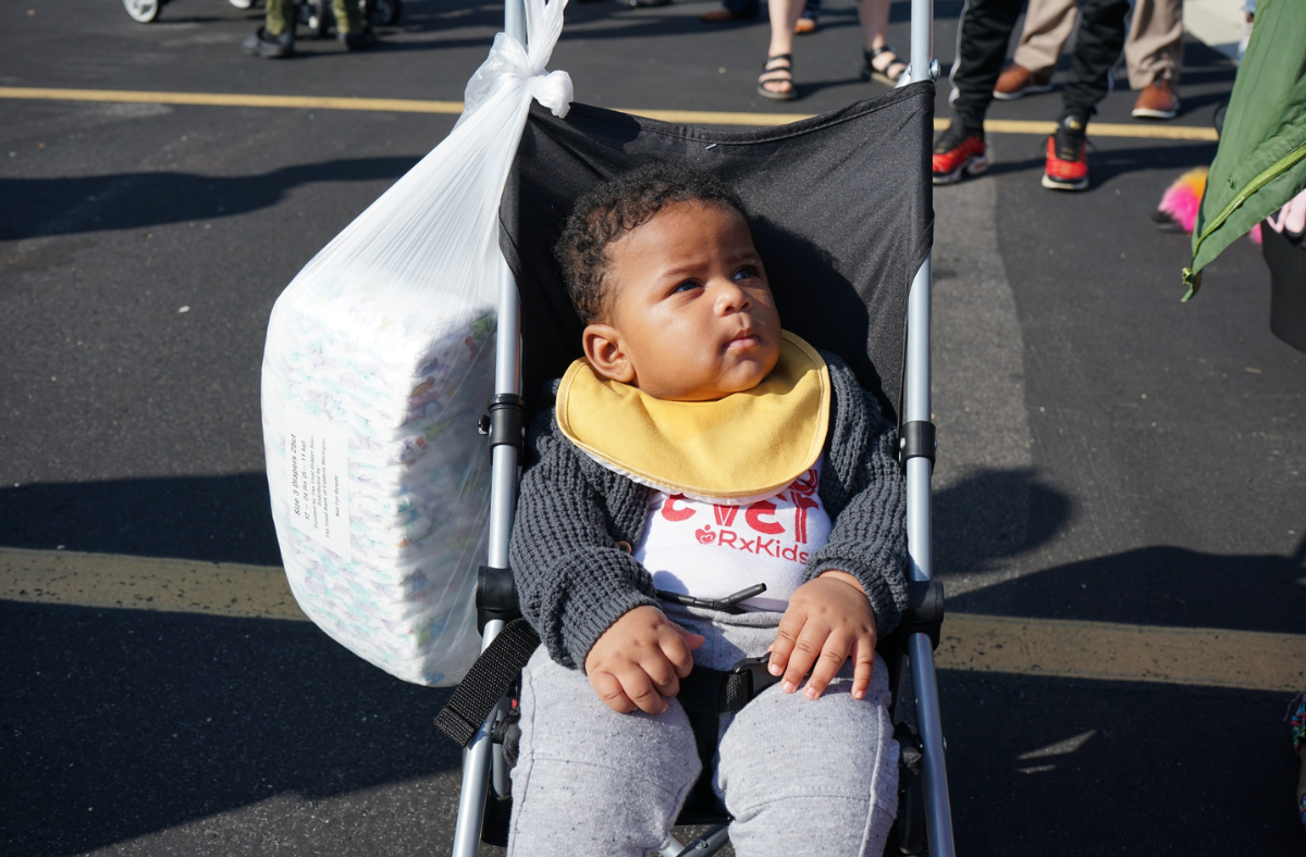 A young child sits in a stroller outdoors, wearing a yellow bib and dark sweater, with a bag of diapers hanging from the stroller handle. Several people stand in the background on a sunny day.