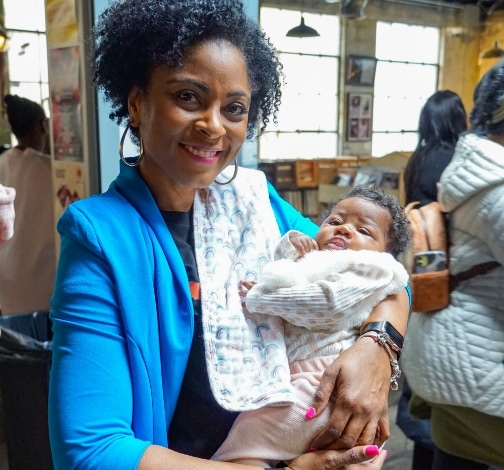 A smiling woman in a blue jacket holds a baby wearing a light-colored outfit and patterned bib in a bright indoor setting, with other people and large windows in the background.
