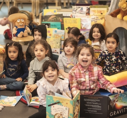 A group of young children sit together on the floor in a classroom, smiling and holding open books, surrounded by colorful books and stuffed animals.