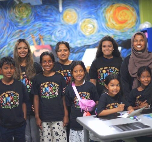A group of smiling children and adults wearing matching "Summer Reading 2024" T-shirts pose together in a colorful classroom with a Starry Night mural in the background.