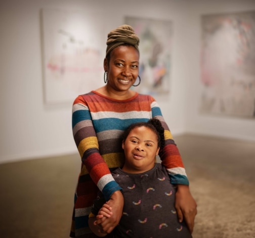 Two women smile together in an art gallery. One stands behind the other, resting her hands on her shoulders. Both wear colorful, patterned clothing. Abstract paintings hang on the walls in the background.