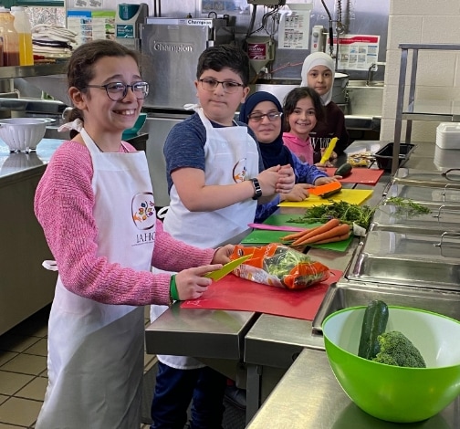 Five children wearing aprons are standing in a kitchen, smiling and preparing vegetables such as carrots, broccoli, and cucumbers on cutting boards. There are bowls and kitchen supplies on the counters.