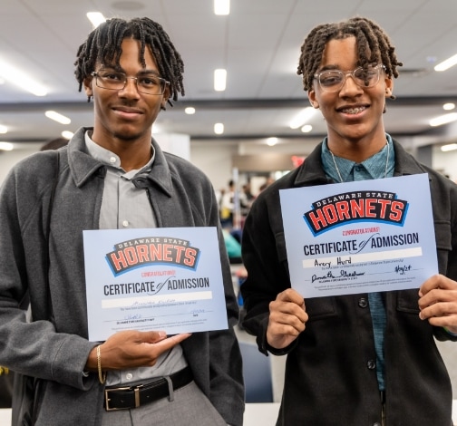 Two young men smile and hold up Delaware State Hornets certificates of admission in a brightly lit room, celebrating their acceptance to the university.