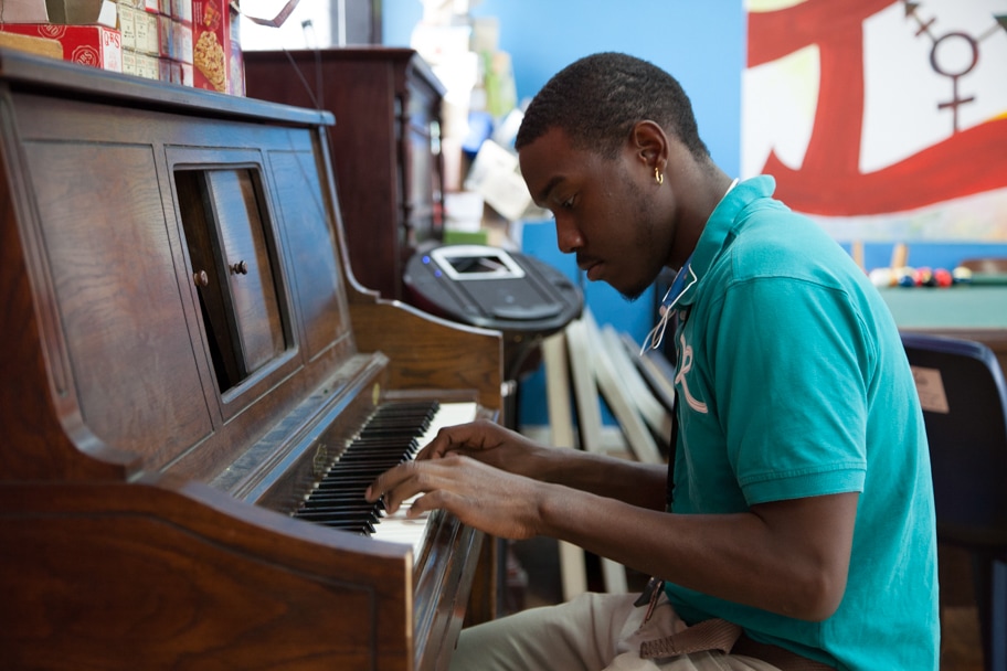 A young man wearing a teal shirt plays an upright wooden piano intently in a colorful room with artwork, books, and a music player in the background.