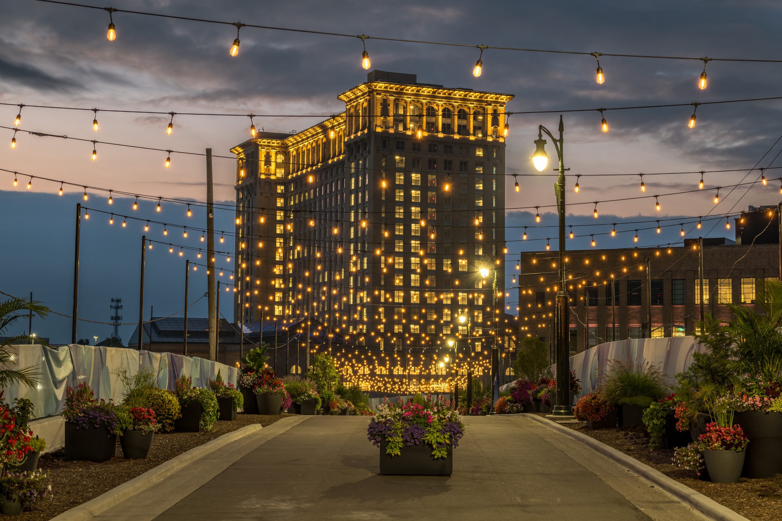 Tall building lit with many yellow lights at dusk; string lights hang above a pathway lined with potted plants and flowers, creating a festive atmosphere against a cloudy evening sky.