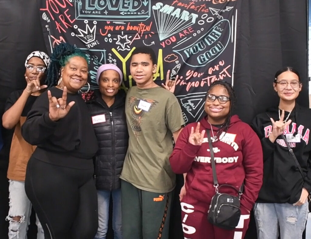 Six people smile and pose in front of a colorful, inspirational backdrop. Several are making the "I love you" hand sign in American Sign Language. The backdrop features positive messages and doodles.