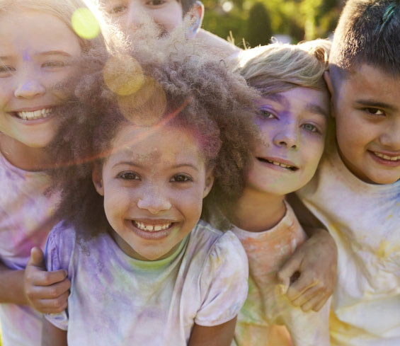 A group of five smiling children with colorful powder on their faces and clothes stand close together outdoors, enjoying a sunny day. Sunlight creates a lens flare over the scene.
