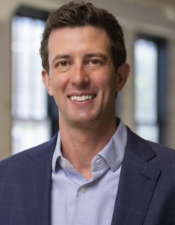 A man with short dark hair, wearing a blue blazer and light blue collared shirt, smiles at the camera. The background shows blurred windows and an indoor setting.
