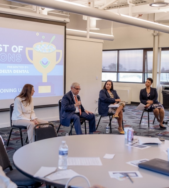 Four people are seated in a panel discussion in front of a screen displaying “Best of Champions presented by Delta Dental.” A round table with papers, a water bottle, and a can is in the foreground.