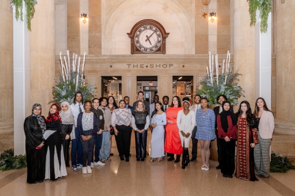 A diverse group of people pose together for a group photo in an elegant lobby with tall columns, floral arrangements, and a large clock above a sign that reads "THE SHOP.