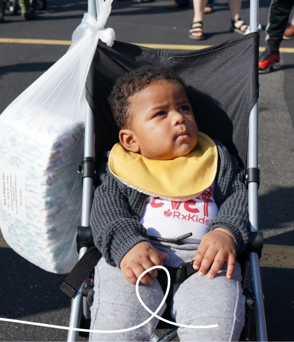 A baby sits in a stroller wearing a yellow bib and gray sweater. A large bag of diapers hangs from the stroller handle. The scene appears to be outdoors with people standing in the background.