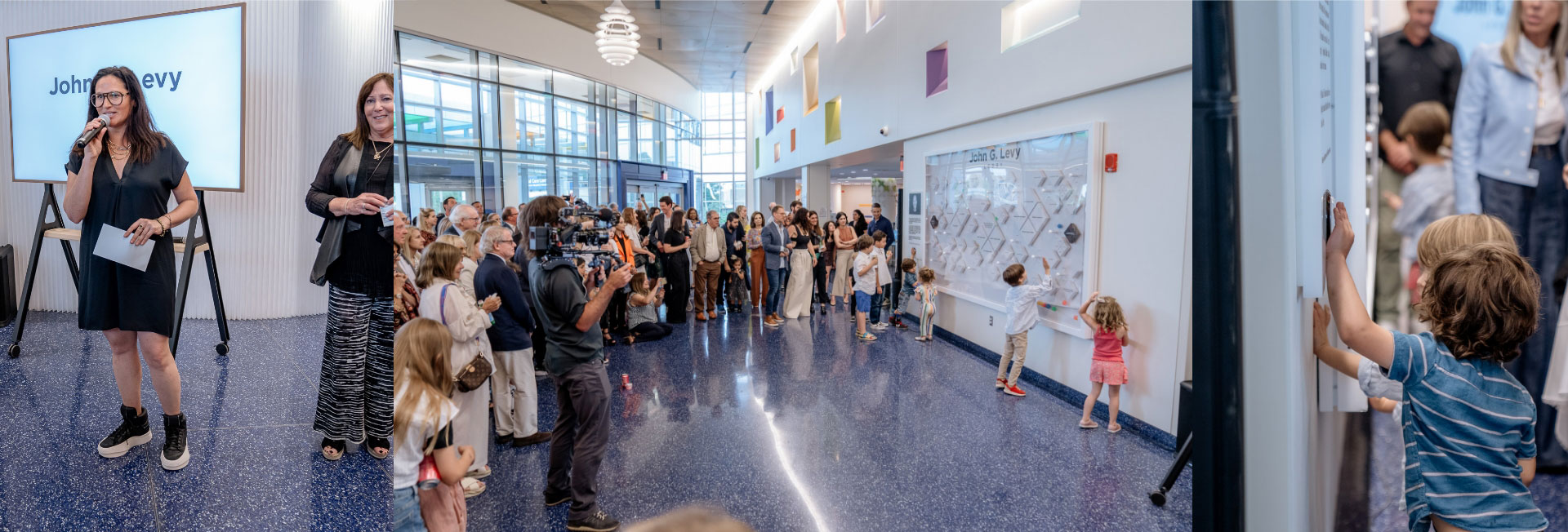 A woman speaks into a microphone at an indoor event while a crowd listens, photographers take pictures, and children interact with a wall display in a bright modern hallway.