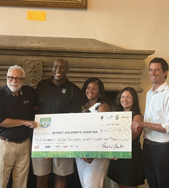 Five people stand indoors, smiling and holding a large ceremonial check for $407,098.00 made out to Detroit Children's Charities. A fireplace with a decorative face sculpture is in the background.