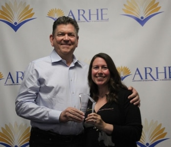 Two people smiling and holding glass awards in front of a backdrop with the ARHE logo, featuring a stylized book and sun design.