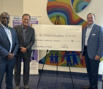 Three men in suits stand smiling beside a large check for $125,000 made out to The Children’s Foundation. A colorful mural and a hospital sign are visible in the background.