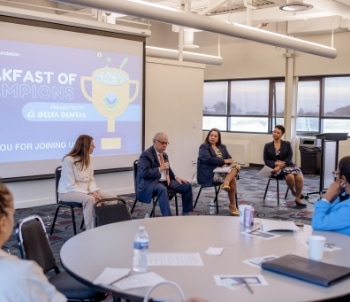Four people sit at the front of a conference room, participating in a panel discussion. A screen behind them reads "Breakfast of Champions presented by Delta Dental." Attendees are seated at round tables.